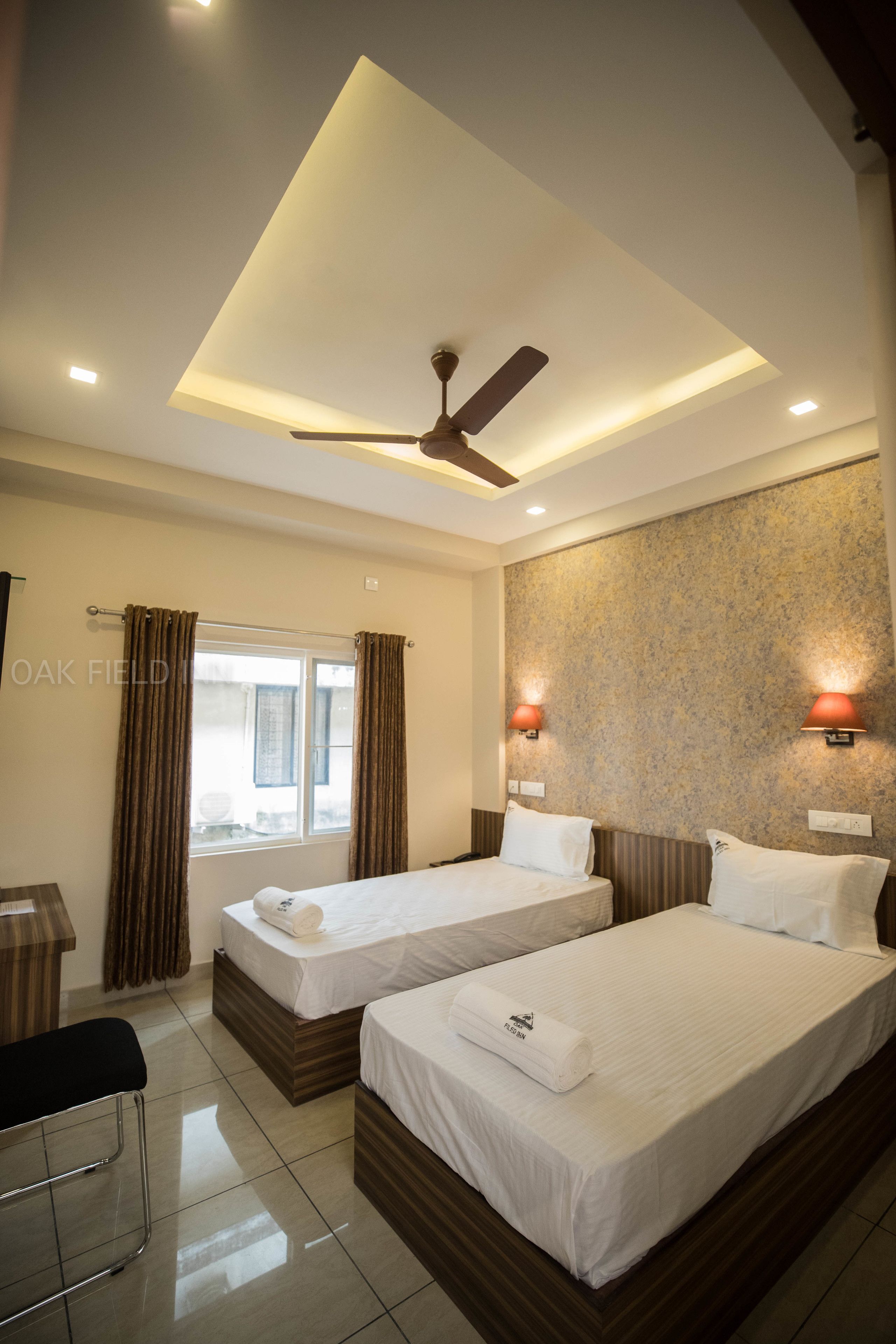 A hotel room with two beds, a ceiling fan, a window with curtains, and wall-mounted lights.