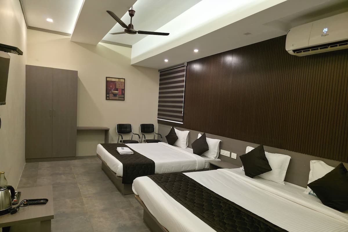 A hotel room with two beds, a ceiling fan, an air conditioner, a desk with a kettle and remote control, and a wardrobe.