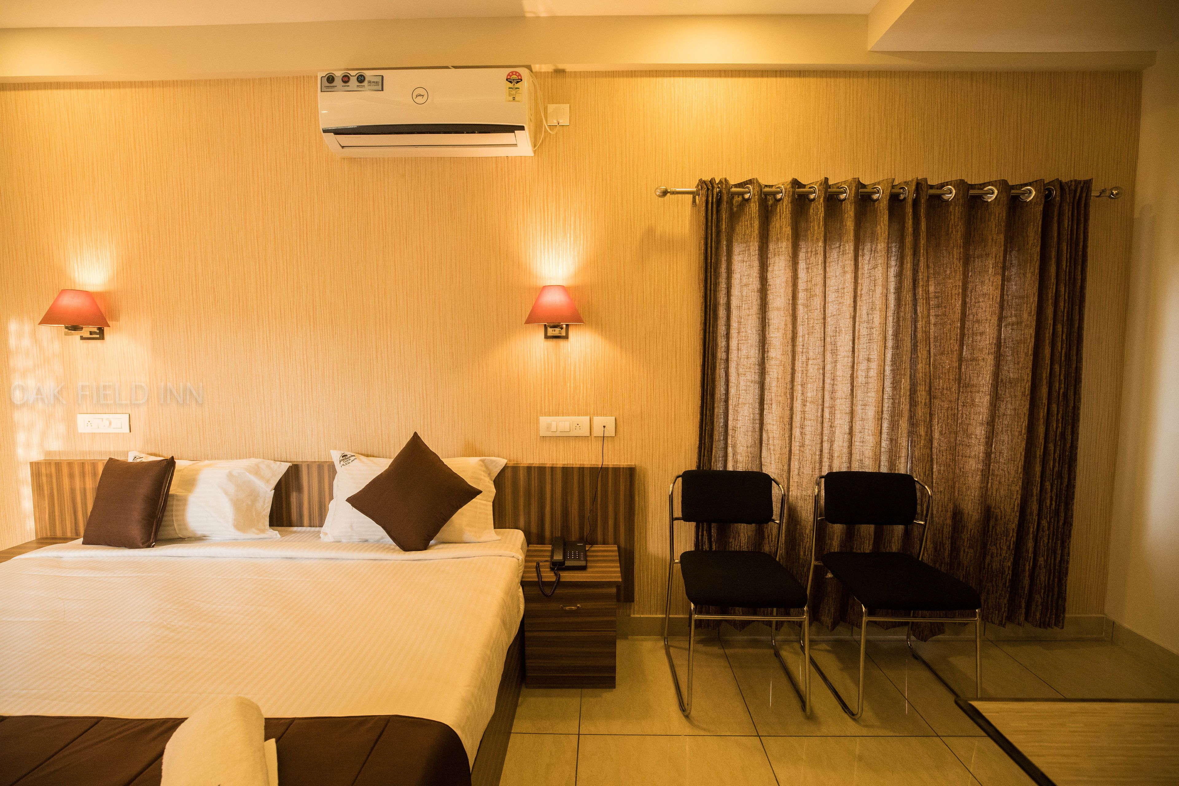 Een hotelkamer met een bed, nachtlampjes, een telefoon, twee stoelen en een airconditioner.