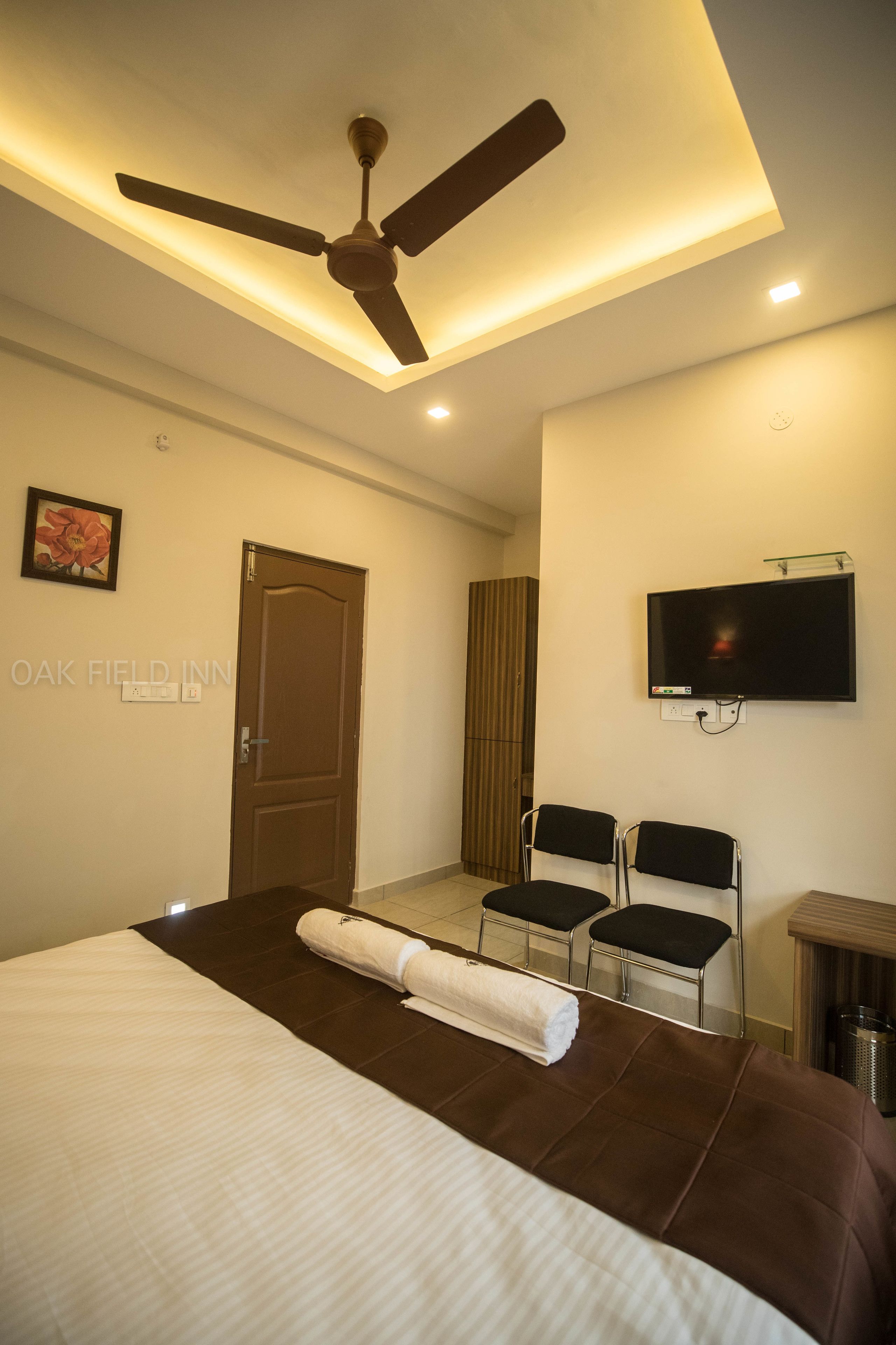 Quarto de hotel com cama, ventilador de teto, TV fixada na parede e duas cadeiras.