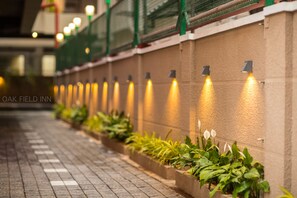 Un chemin bordé de jardinières et illuminé par des lampes fixées aux murs, menant à l’entrée d’un bâtiment.