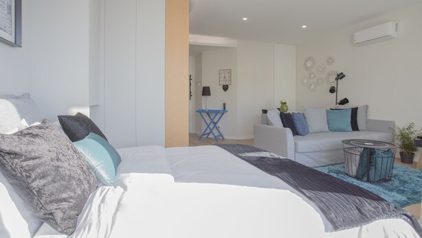 A modern, well-lit bedroom with a large bed, a small side table, and a sofa in the adjacent living area. - Liiiving | Bright Light Studio (Porto)