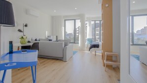 A modern living room with a grey sofa, a blue chair, and a small wooden table. There is a large window providing a view of the city. - Liiiving | Bright Light Studio (Porto)
