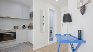 A modern kitchen with white cabinets, a built-in oven, and a microwave. There's a blue table with a lamp and a vase on the right. - Liiiving | Bright Light Studio (Porto)