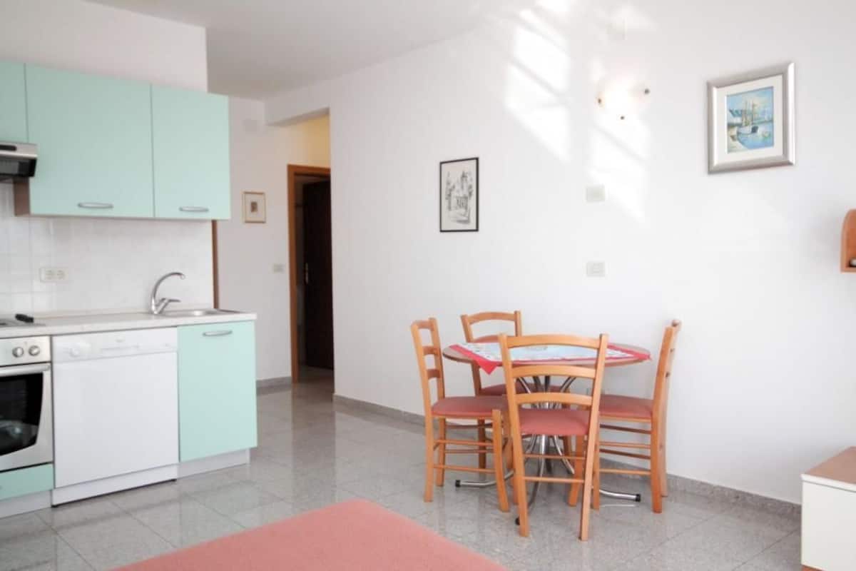 A compact kitchen with teal cabinets, a dishwasher, and a dining area with a wooden table and chairs.