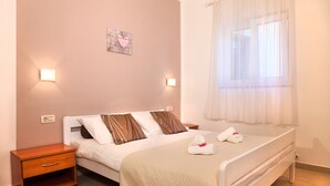 A neatly made bed with a quilted cover, a bedside table with a drawer, a wall-mounted light, and a window with sheer curtains. - Villa Lidija (Lovran)