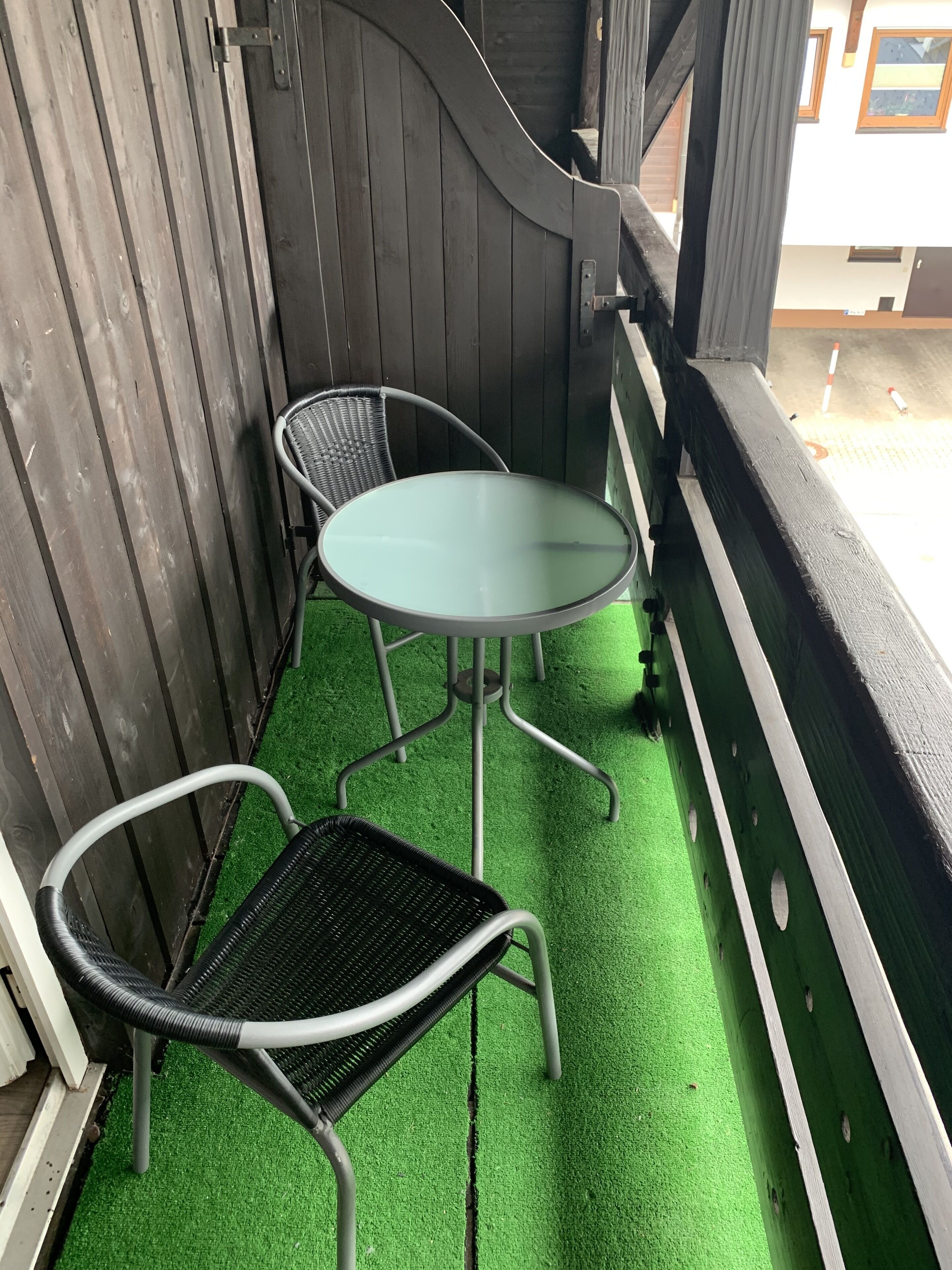 A balcony with a green artificial turf floor, a metal table with a glass top, and two metal chairs.