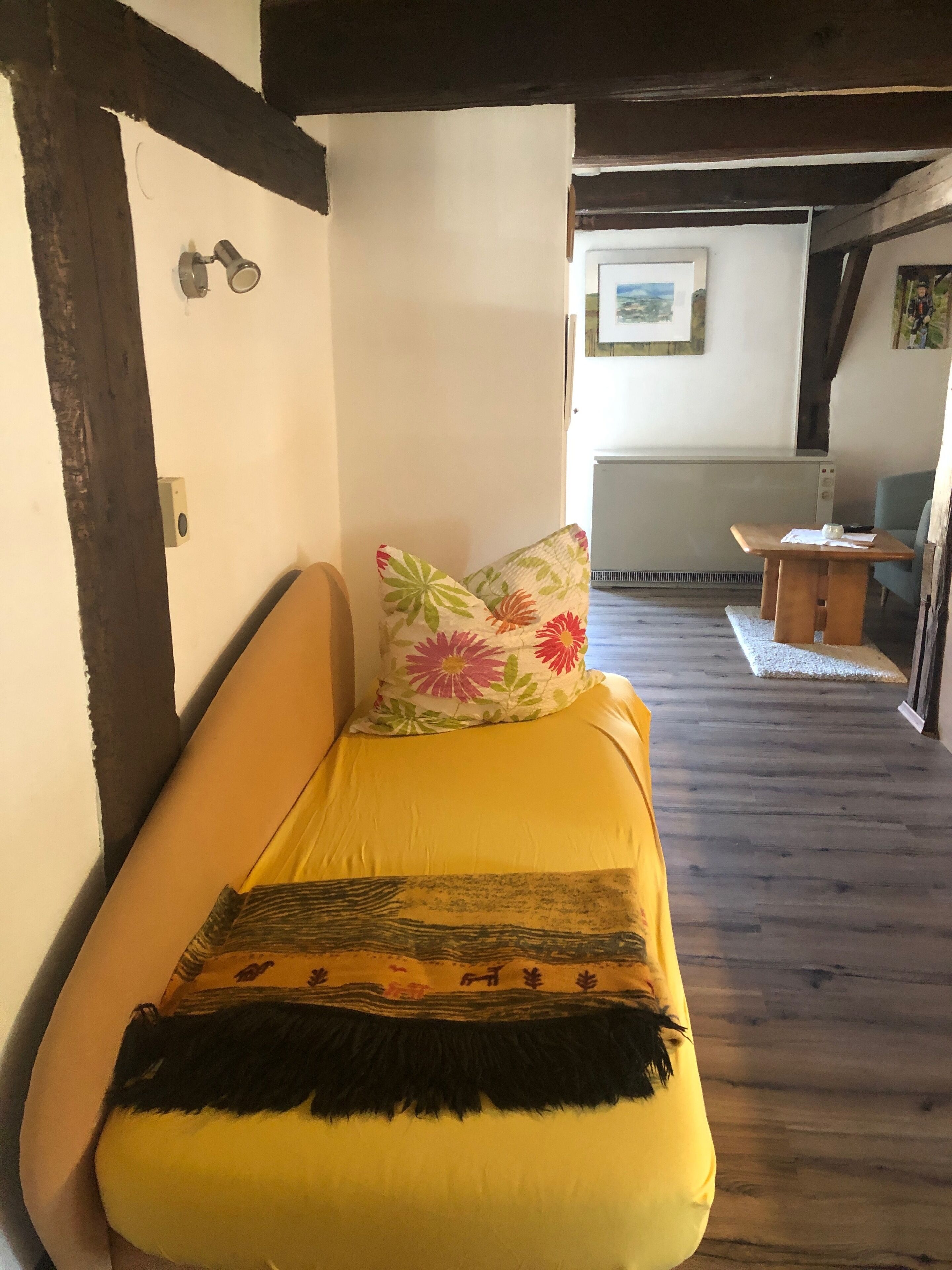 Een kamer met een gele bank, een bloemenkussen en een decoratieve plaid. Er ligt een houten vloer en is een witte muur met een fotolijst.