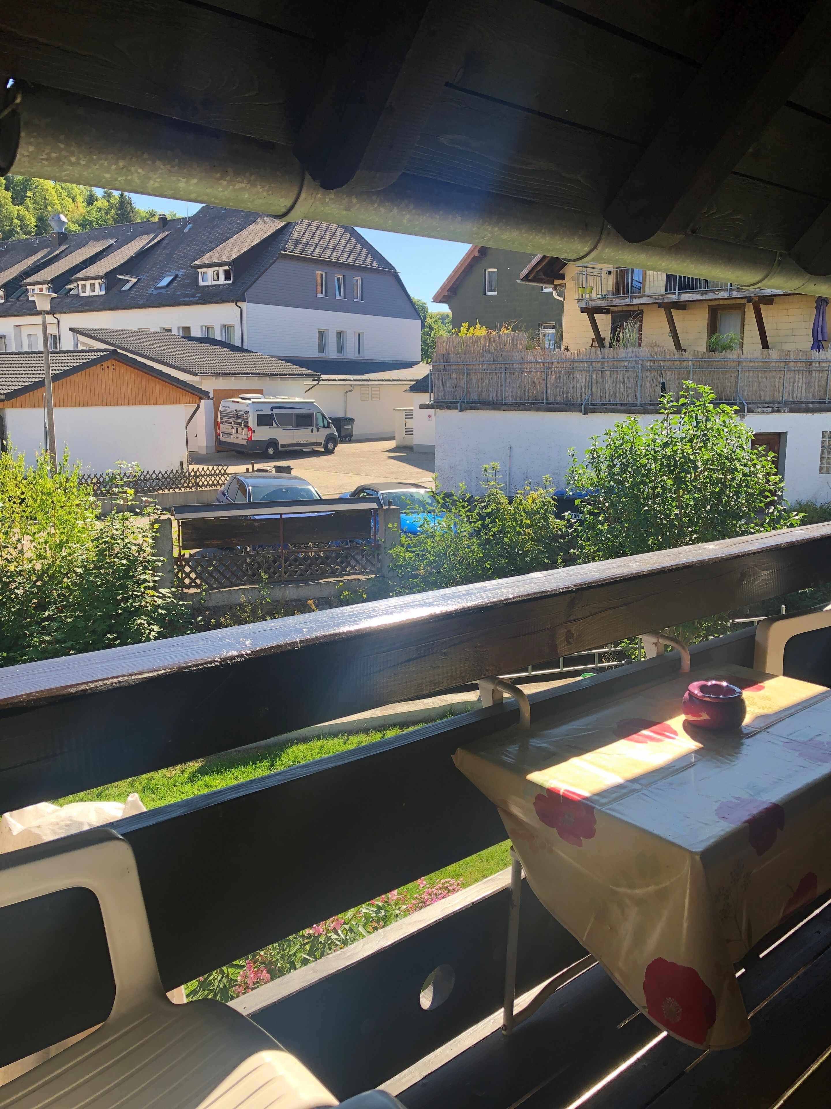 A balcony with a table and a chair, overlooking a residential area with houses and parked cars.