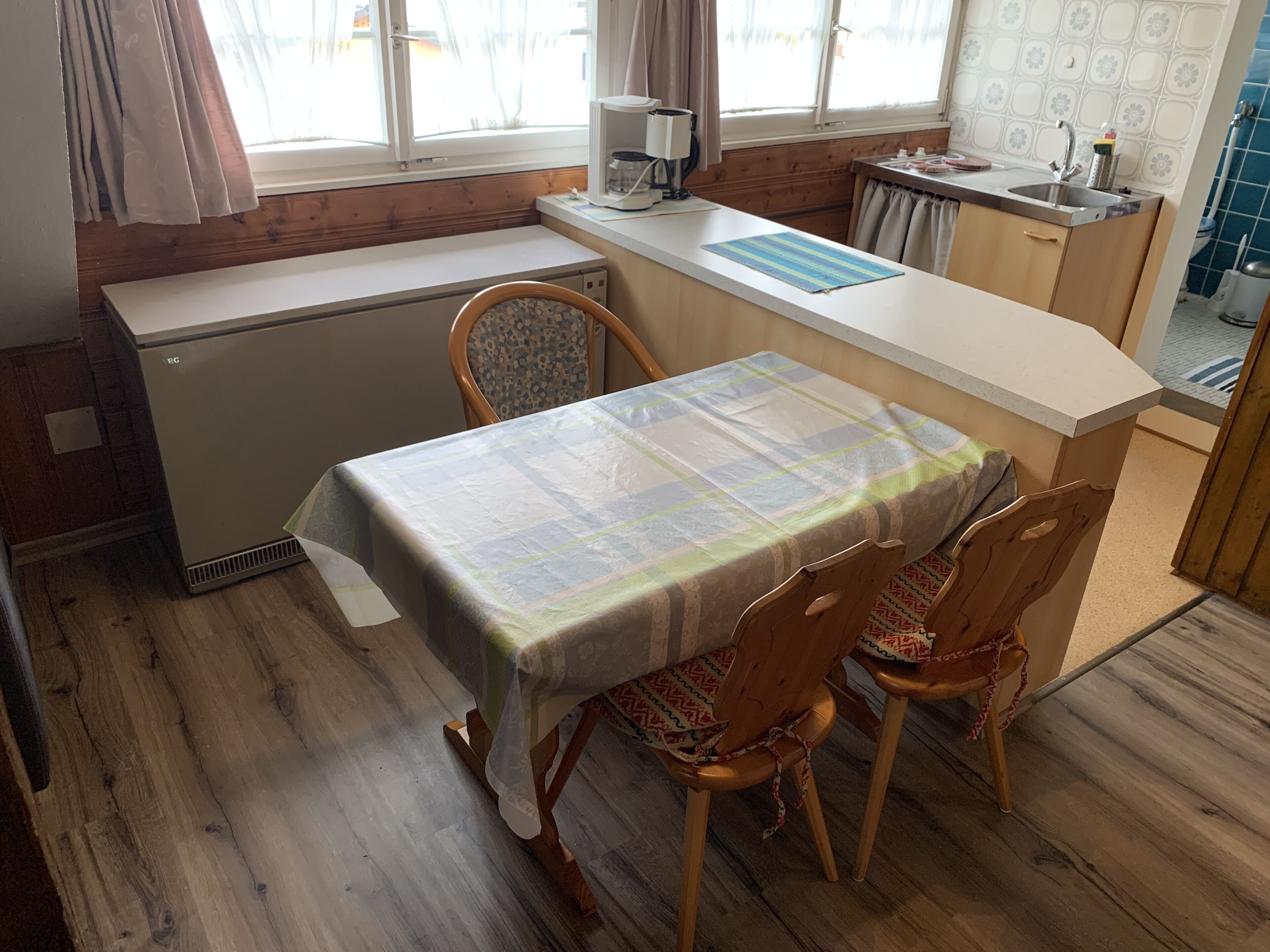 A compact kitchen with a small refrigerator, a dining table with chairs, and a window with curtains.