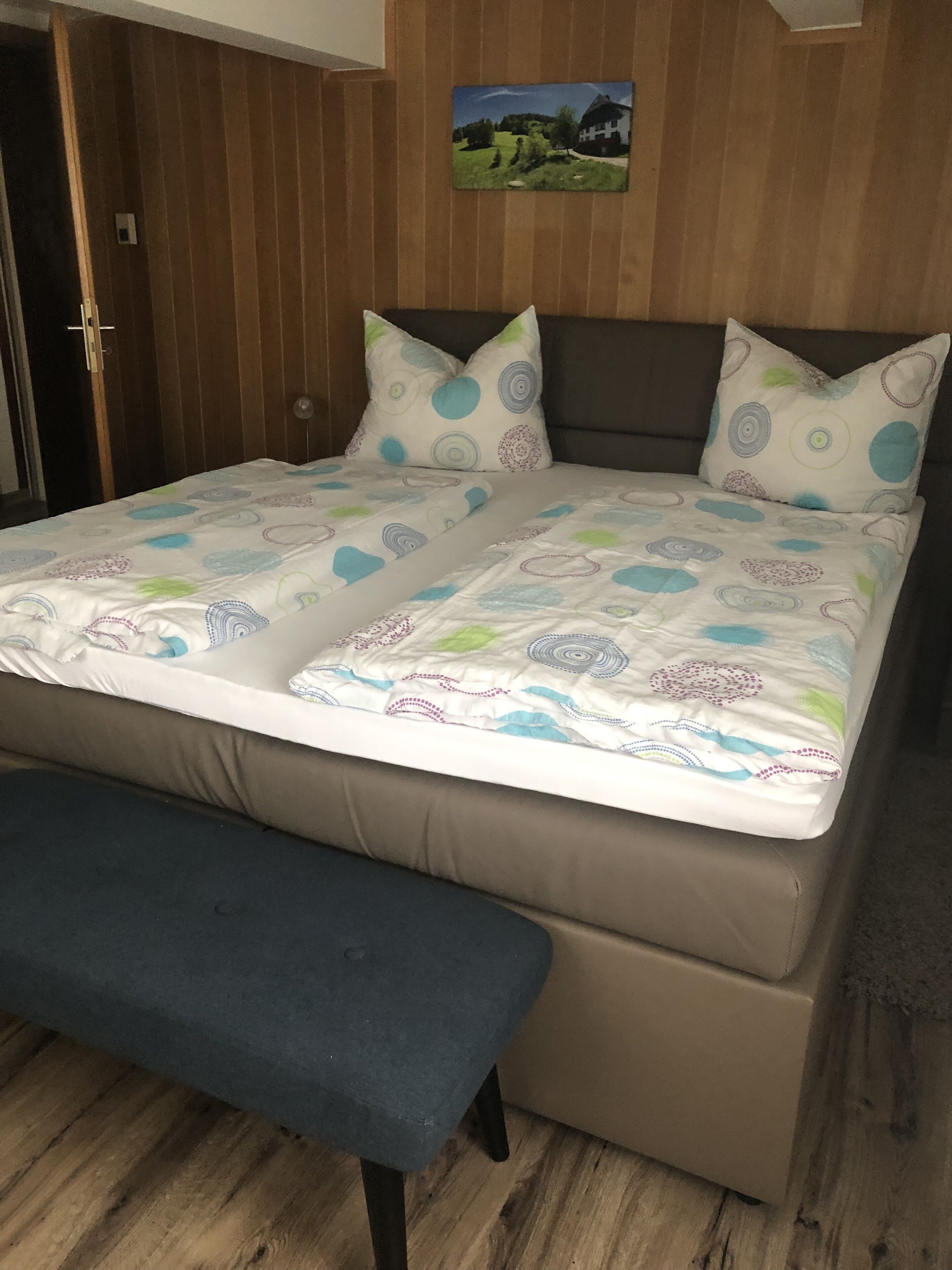Ein Bett mit gemustertem Überwurf, zwei Kissen und eine Bank in einem Zimmer mit Holzvertäfelung und einem Bild an der Wand.