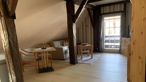 Een gezellige kamer met een houten plafond, een bank, een tafel met een mand, en uitzicht naar buiten door een raam.