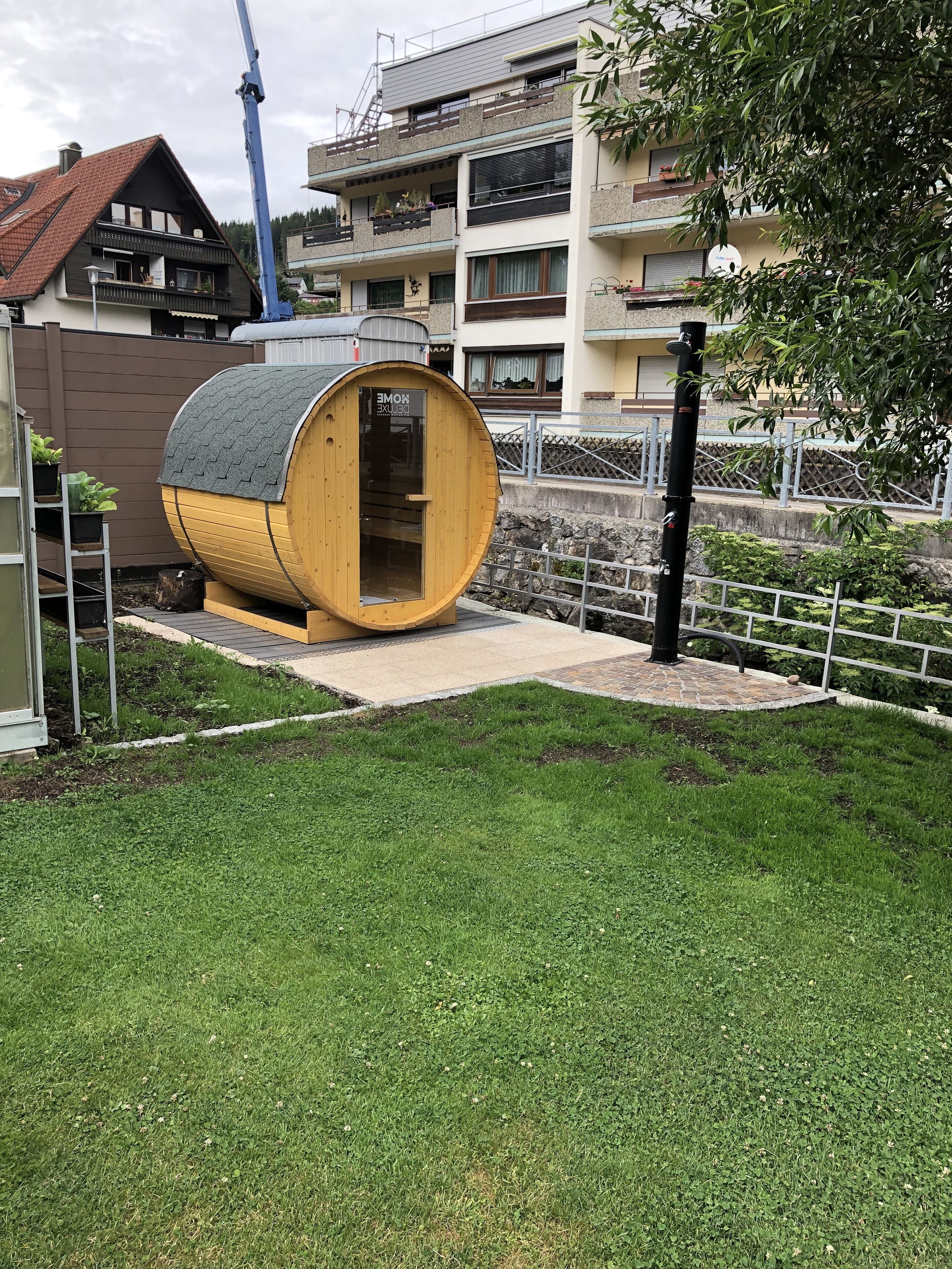 A wooden sauna structure in a garden area with a paved walkway, surrounded by a fence and greenery.