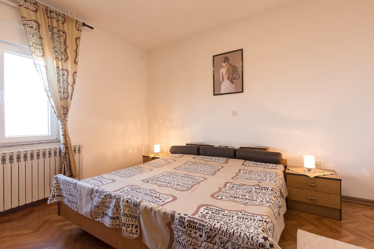 A bedroom with a bed, two bedside tables, a nightstand, a window with curtains, a radiator, and a framed picture on the wall.