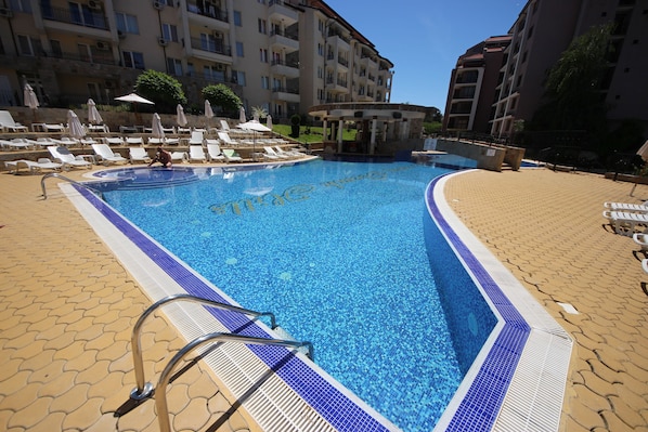 A swimming pool with a blue tiled edge, surrounded by lounge chairs and umbrellas, with a building in the background.