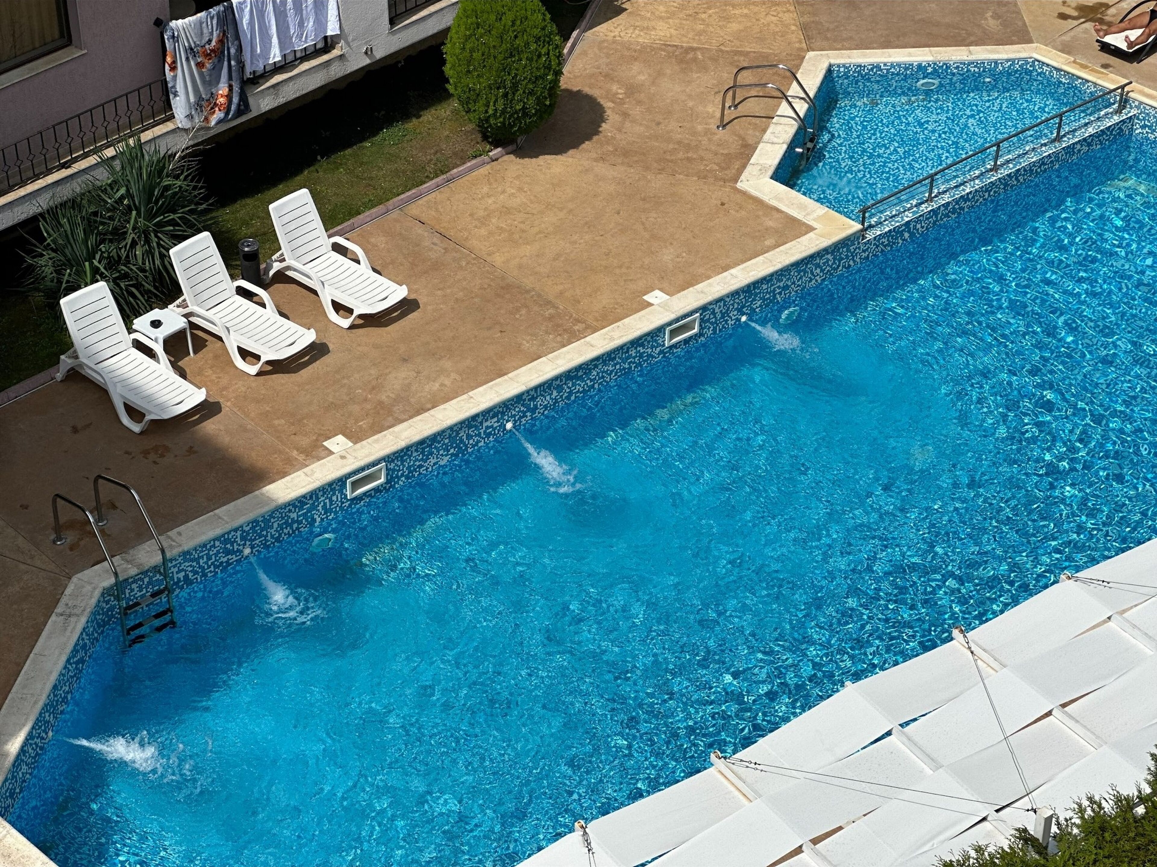 A rectangular swimming pool with a tiled edge, surrounded by white lounge chairs and a small garden area.