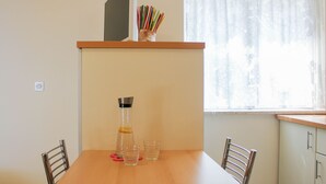 Un pequeño comedor con una mesa de madera, dos sillas, una botella de agua y una taza. Hay una ventana con cortina floral y un estante sobre la mesa que contiene una planta y un florero con bolígrafos.