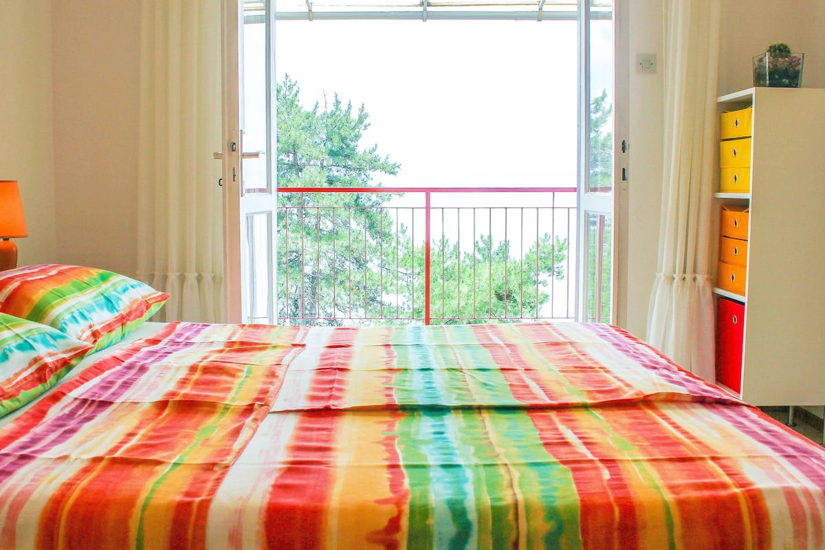 A bedroom with a balcony, a bed with colorful bedding, a lamp, and a dresser with drawers.