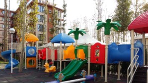 A playground with various play structures, including slides and swings, surrounded by trees and a building in the background. - Menada Apartments in Tarsis Nova Complex (Sunny Beach)
