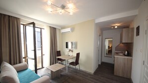 A modern living room with a sofa, a dining table, and chairs. There is a TV on the wall and an air conditioning unit. The room has wooden flooring and a large window with curtains. - Menada Apartments in Tarsis Nova Complex (Sunny Beach)