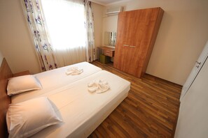Quarto de hotel com piso de madeira, cama com lençóis brancos, guarda-roupa de madeira, escrivaninha com espelho e janela com cortinas florais.