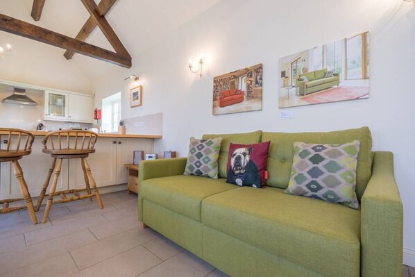A green sofa with patterned cushions, a wooden dining table with chairs, and a kitchen area with white cabinets and a red fire extinguisher.