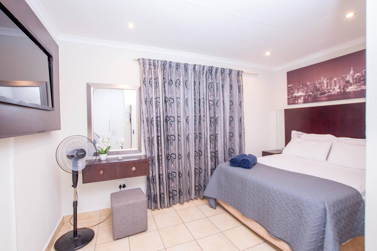 A hotel room with a bed, a bedside table, a mirror, a standing fan, and a curtain with a cityscape print.