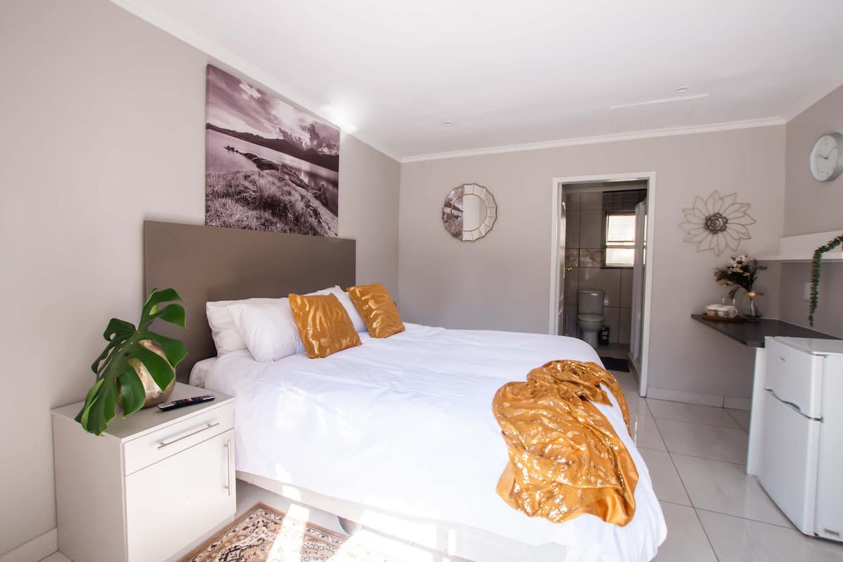 A bedroom with a bed, bedside table, and a plant. There is a door leading to a bathroom and a kitchenette.