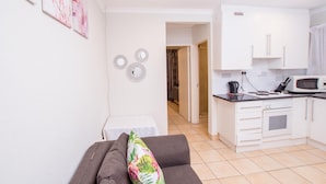 A modern living room with a grey sofa and a colorful pillow, a kitchen with white cabinets and a microwave, and a hallway leading to another room. - Prestigious Apartments (Randburg)