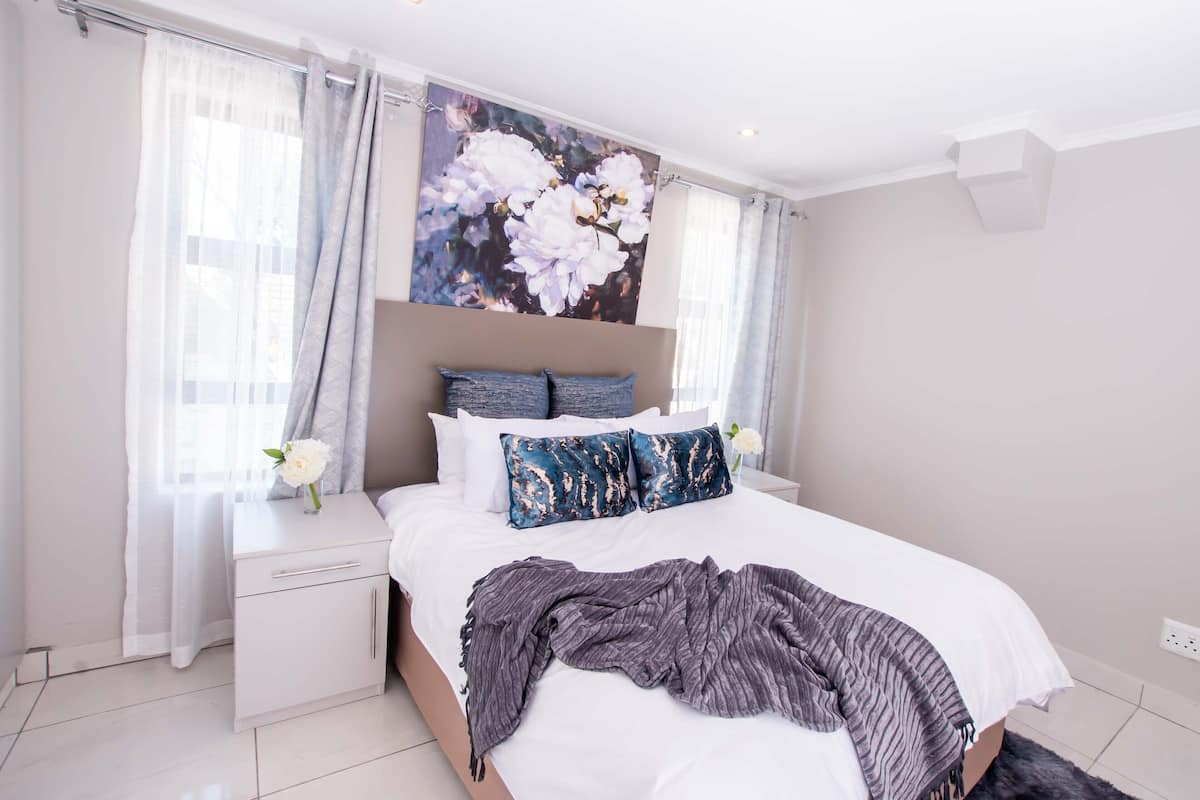 A bedroom with a bed, white nightstands, a large floral painting, and sheer curtains.