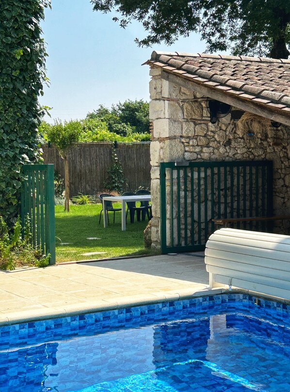 A swimming pool with a stone wall and a wooden deck. There is a table and chairs in the background.