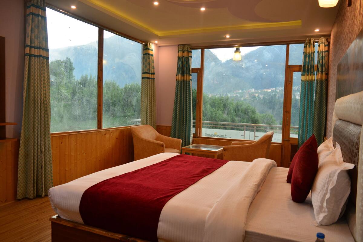 A hotel room with a large bed, a wooden headboard, a small table, and a view of mountains through large windows.