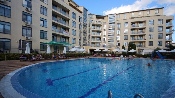 A swimming pool surrounded by a multi-story building with balconies, some with air conditioning units. - Menada Apartments in Rainbow 1 Complex (Sunny Beach)