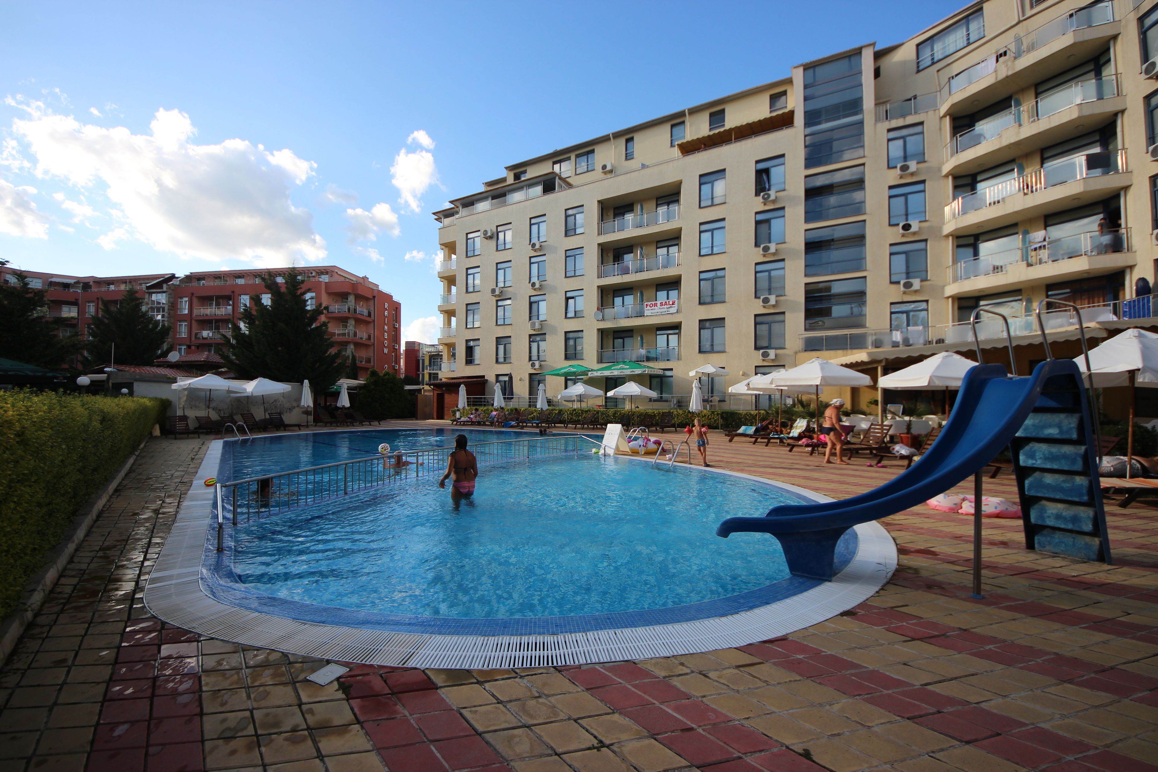 A swimming pool with a slide, surrounded by lounge chairs and umbrellas, with apartment buildings in the background.