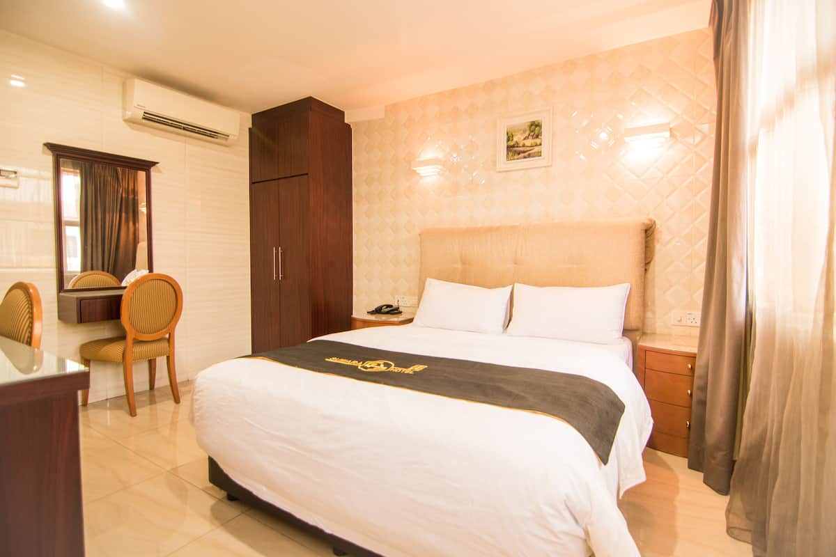 A hotel room with a large bed, a desk with a chair, a wardrobe, and a nightstand with a lamp.