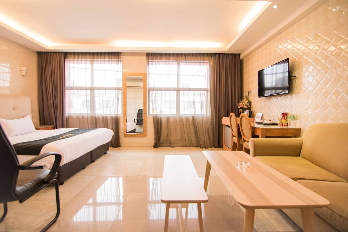 A hotel room with a bed, a desk, a sofa, a TV mounted on the wall, and large windows with curtains.
