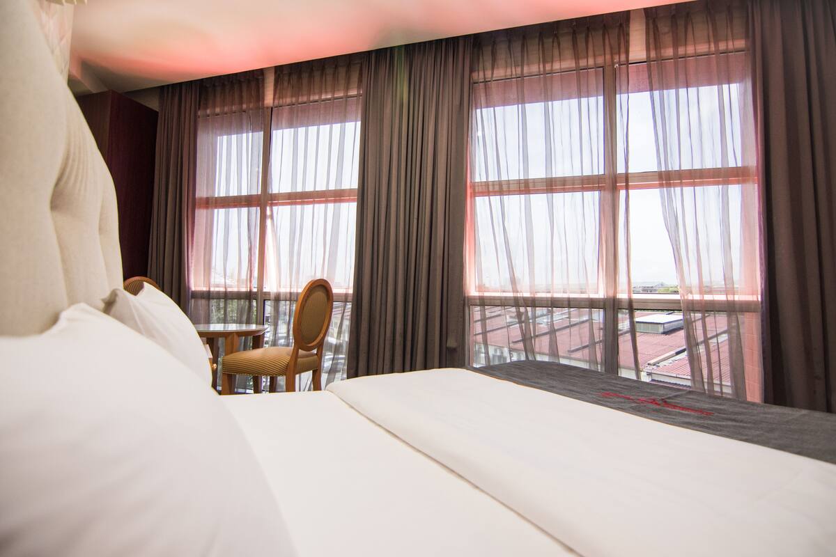 A hotel room with a large bed, a chair, and a view of the outside through sheer curtains.