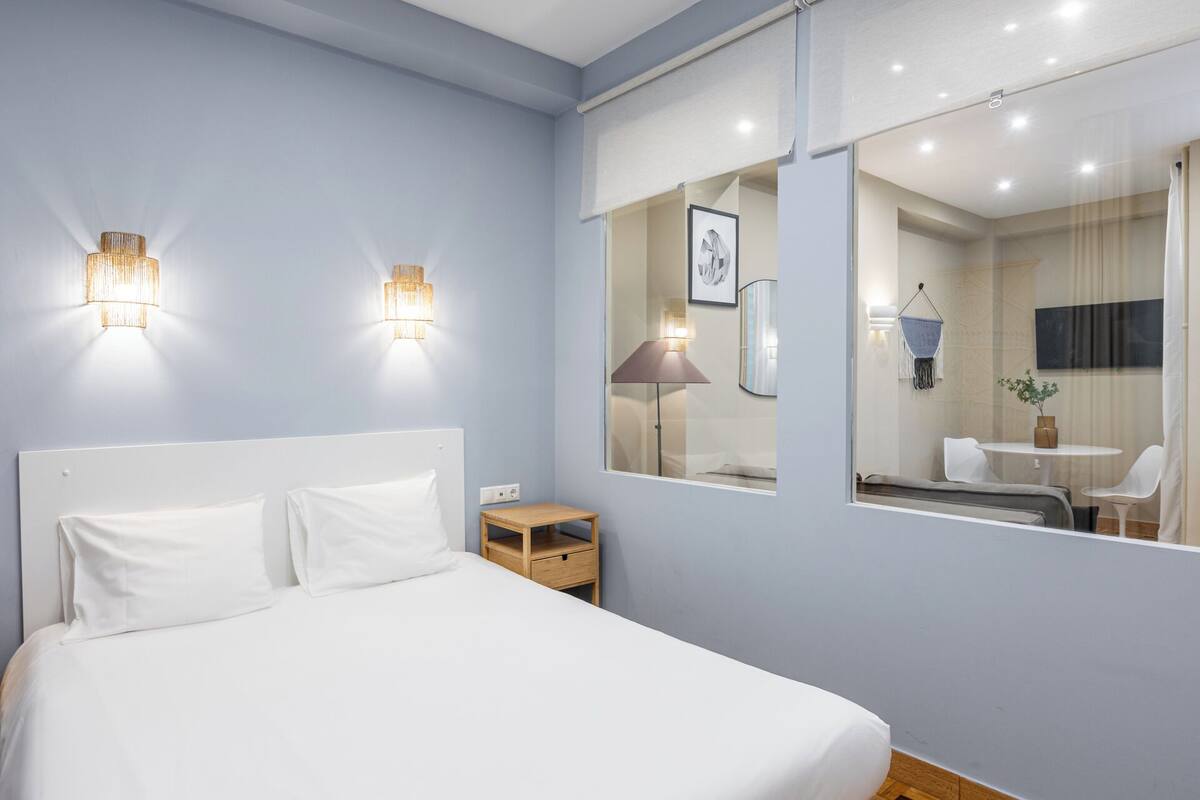 Quarto com uma cama grande, luminárias de cabeceira, mesa de cabeceira e um espelho refletindo um banheiro com pia e cadeira.