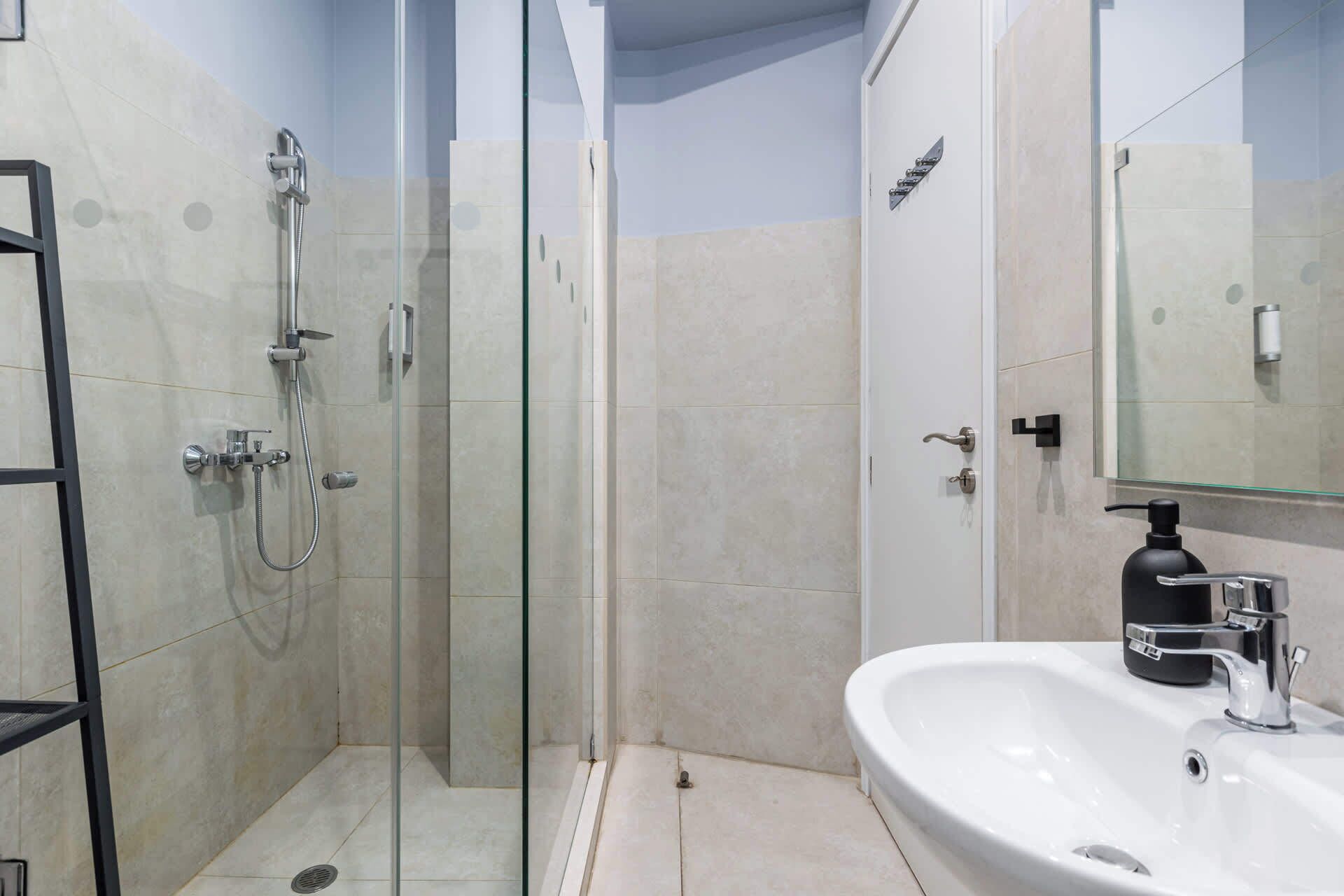 A modern bathroom with a glass-enclosed shower, a white sink with a black soap dispenser, and a mirror above the sink.