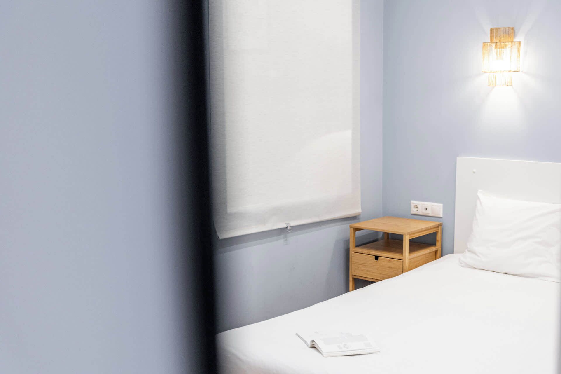 Une chambre d’hôtel comprenant un lit, une table de chevet, une lampe fixée au mur et une fenêtre avec un store.