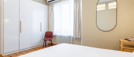 A bedroom with a bed, a wardrobe, a chair, a mirror, and a window with curtains.