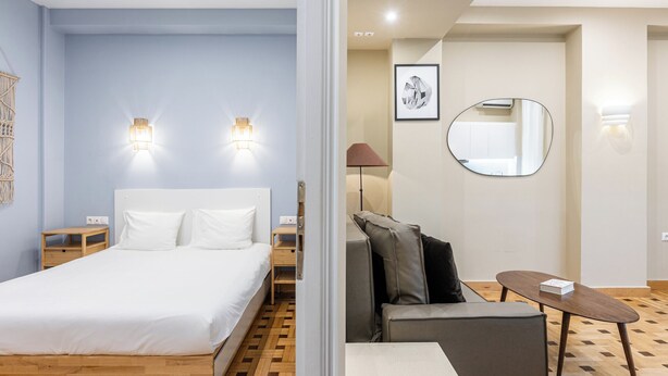 Camera d'albergo moderna con un letto in legno, un divano grigio, un tavolino e uno specchio rotondo.