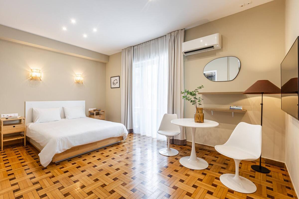 Quarto de hotel com cama, mesas de cabeceira, uma mesa redonda com cadeiras, espelho e ar condicionado.