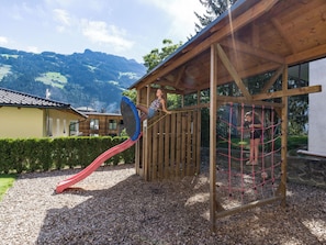 A wooden play structure with a slide and netted climbing area, set against a backdrop of mountains and a house. - Garni Nill (Schwendau)