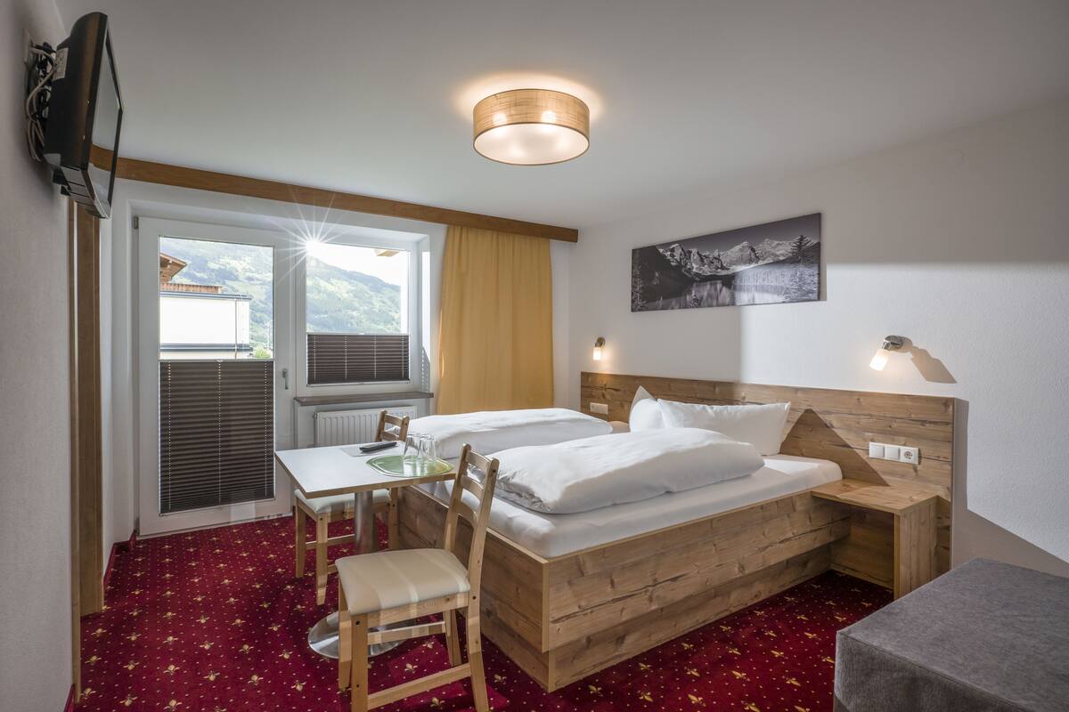 Une chambre d’hôtel avec un lit en bois, un bureau et une chaise. Il y a une fenêtre avec des stores, une lampe et un tableau au mur.