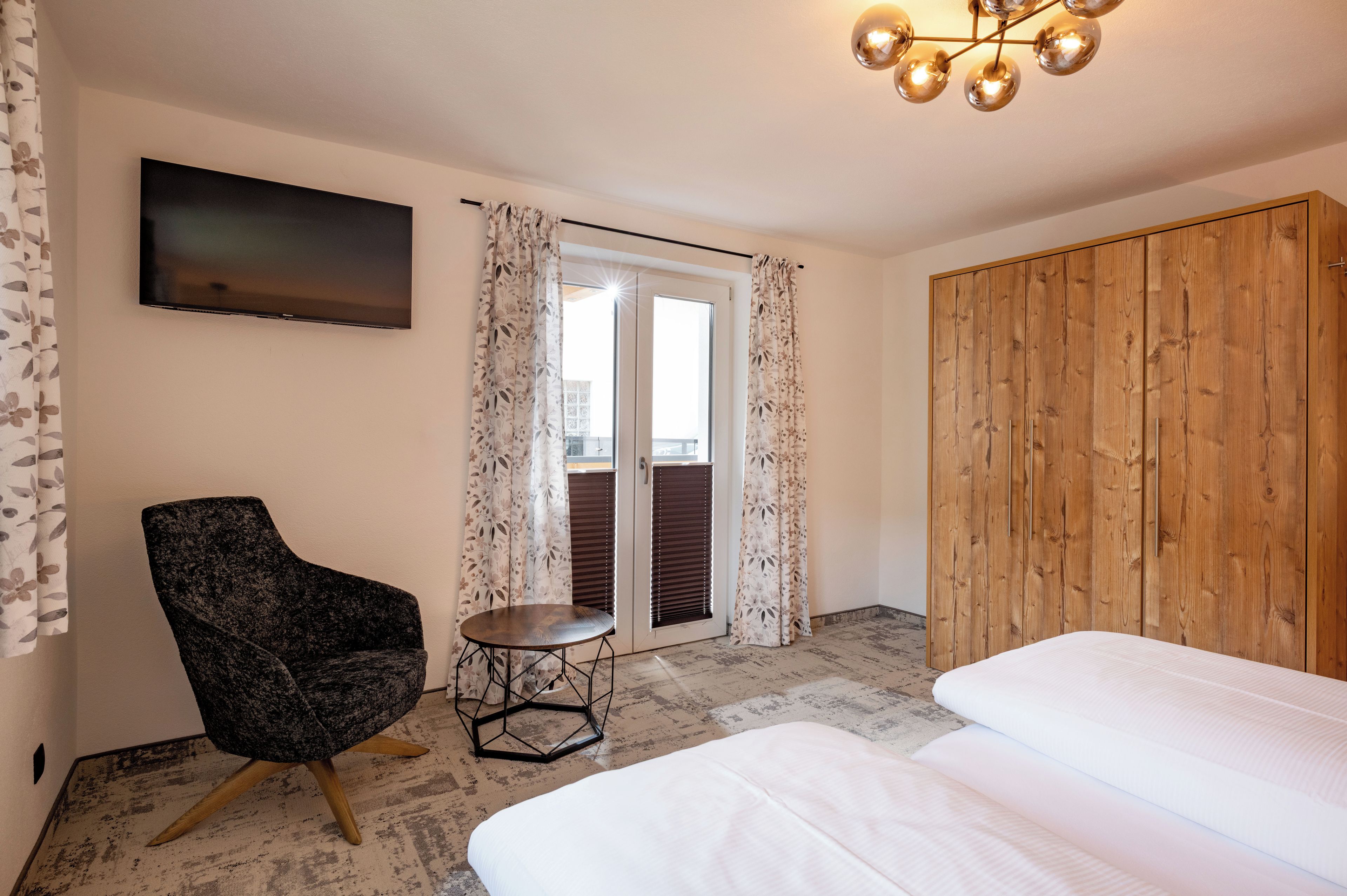 Habitación de hotel con cama, una silla, televisión, armario de madera y balcón con cortinas.