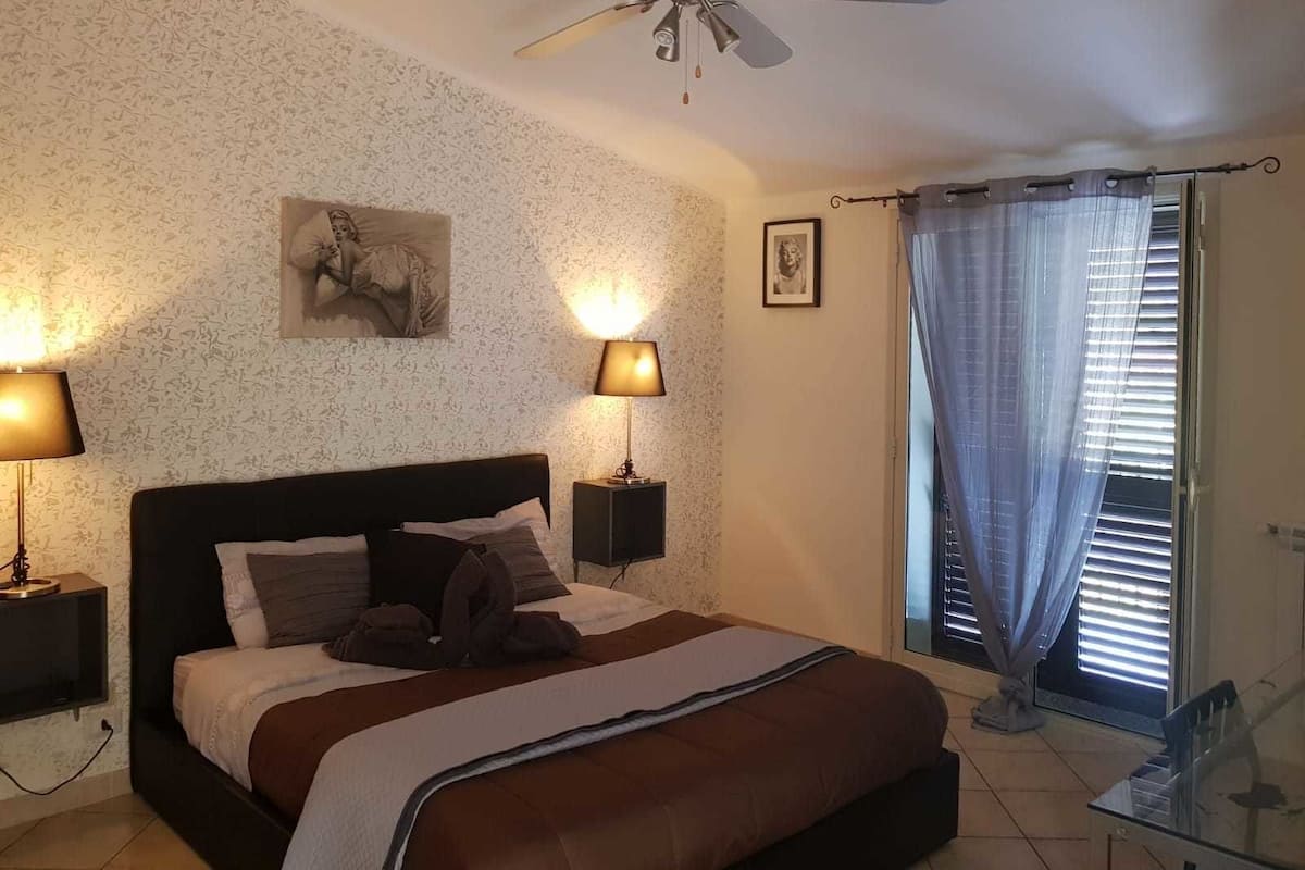 Une chambre à coucher comprenant un lit, deux lampes de chevet, un ventilateur de plafond, une fenêtre avec des rideaux et un mur orné d’un motif floral.