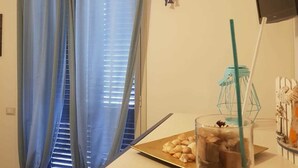 A room with a window covered by blue curtains, a small fish decoration, a TV on the wall, a glass jar with a straw, and a tray with nuts on a table. - B&B Fragranza Di Sicilia (Giardini Naxos)