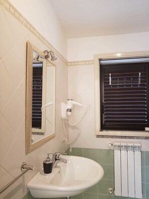 Un baño con un lavamanos blanco, un espejo con marco dorado, una ventana con persianas y un perchero.