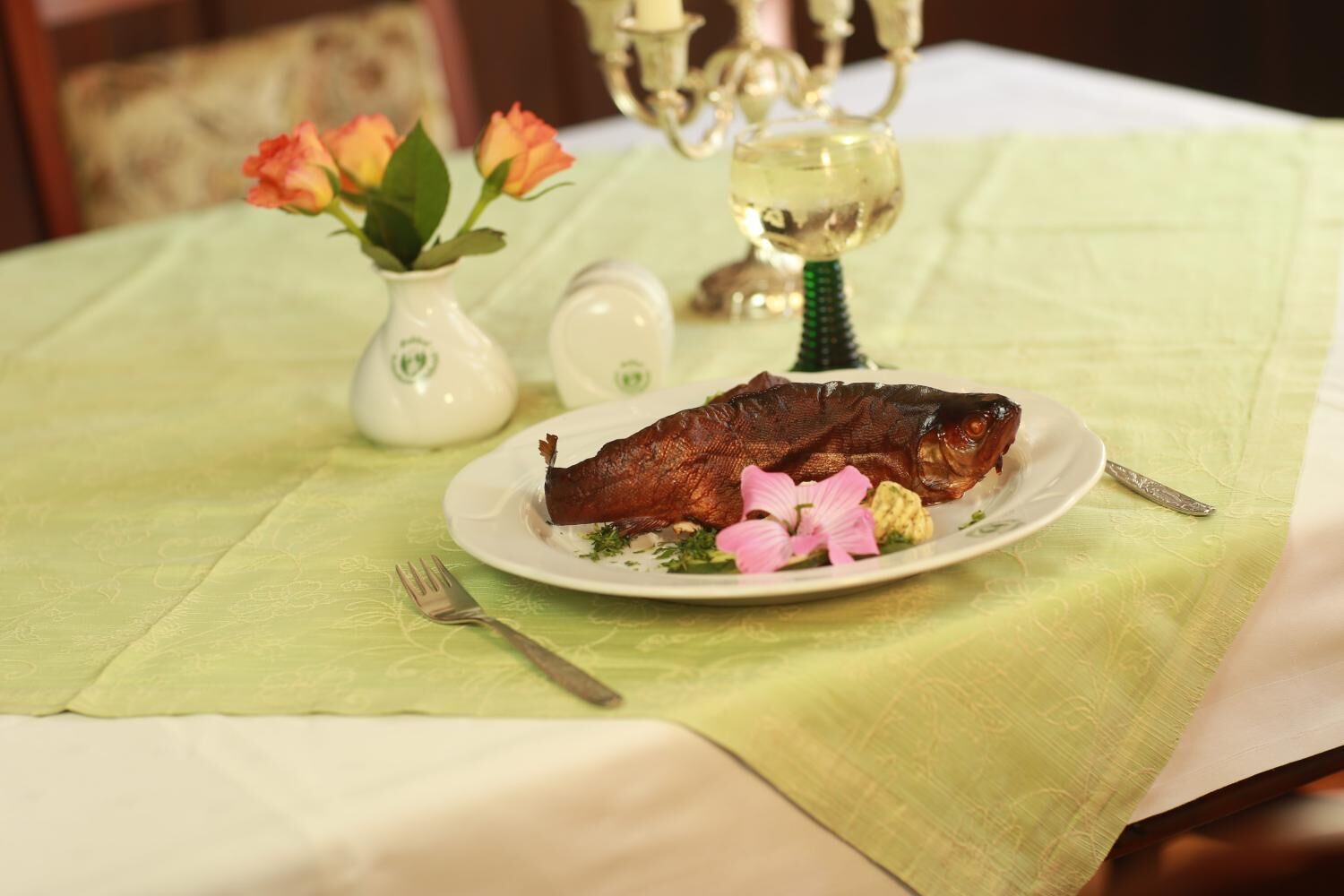 A plate with a cooked fish on a bed with a green tablecloth, a fork, a vase with flowers, and salt and pepper shakers.